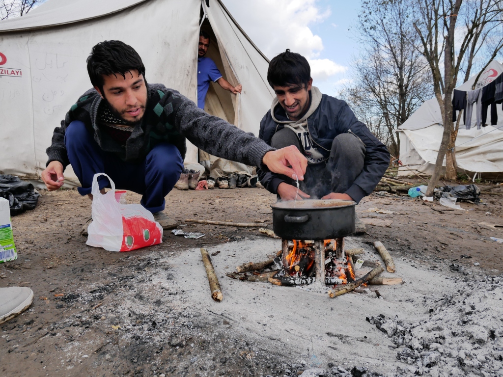 Kaksi afgaanimiestä keittää teetä pienessä kattilassa nuoriolla, taustalla pakolaisteltta.